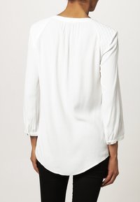 Vit blus med långa ärmar, rynkad textur vid axlarna, rundad nederkant och knäppte manschetter. Slät tyg, inga synliga mönster.