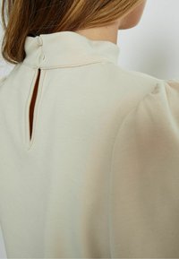 Light beige top featuring a high collar, button closure at the back, and soft, textured fabric with slightly puffed sleeves.