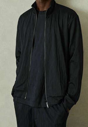 Homme portant une veste zippée noire texturée, une chemise texturée assortie et un pantalon, les mains dans les poches, sur un fond clair.