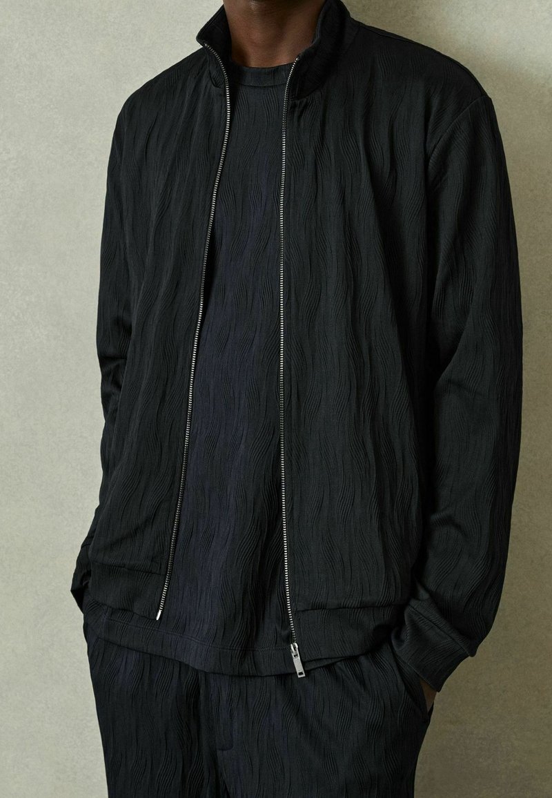 Homme portant une veste zippée noire texturée, une chemise texturée assortie et un pantalon, les mains dans les poches, sur un fond clair.