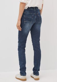 Jeans en denim bleu foncé ajustés, avec une taille mi-haute, deux poches arrière et des détails en tissu texturé. Portés avec des baskets blanches.