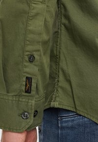 Camicia verde oliva con polsini abbottonati, dettagli sulle cuciture laterali e un'etichetta in tessuto. Realizzata in un misto cotone liscio e testurizzato.