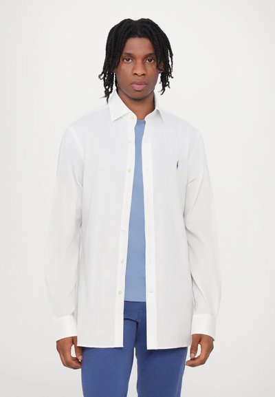 Camicia bianca a maniche lunghe con colletto, abbinata a una t-shirt azzurra chiara con un logo discreto sul petto.