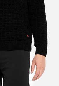 Maglione nero con texture e motivo a griglia, polsini a coste e una piccola etichetta rossa di lato, abbinato a pantaloni neri.