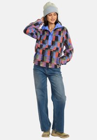 Fleece-pullover med et farverigt ternet mønster i rød, blå, grøn og sort; har en blå krave og forlommer. Bæres med blå jeans.