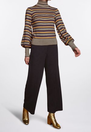 Personne portant un pull à col roulé rayé avec des manches ballon, un pantalon large noir et des bottines dorées brillantes, debout sur un fond blanc.