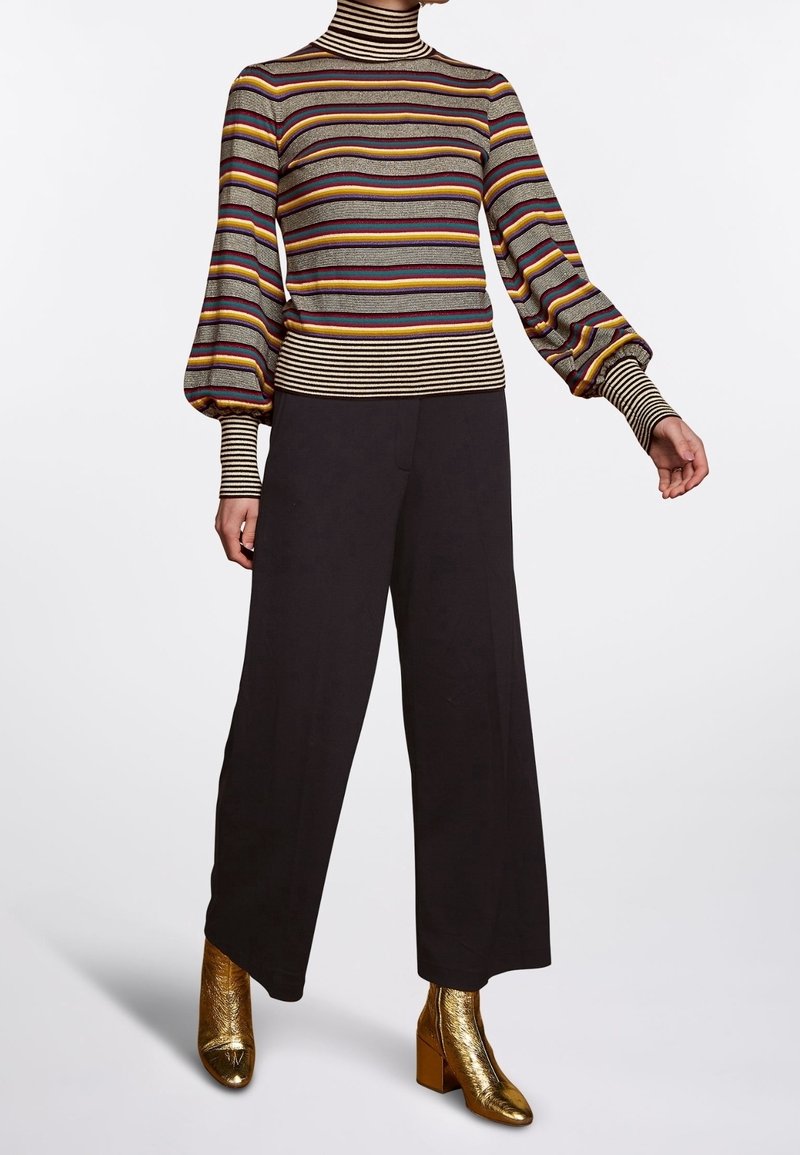 Personne portant un pull à col roulé rayé avec des manches ballon, un pantalon large noir et des bottines dorées brillantes, debout sur un fond blanc.