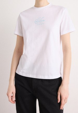 Osoba ubrana w białą koszulkę Calvin Klein z jasnoniebieskim logo, w połączeniu z czarnymi spodniami, stoi na jednolitym tle.