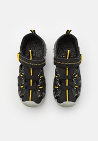 Pari musta ja keltainen lasten sandaaleja säädettävillä remmeillä, suljetuilla varpailla ja teksturoiduilla harmailla pohjilla, kuvattuna ylhäältä valkoisella taustalla.