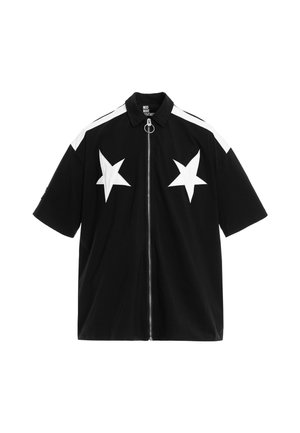 Persona joven que lleva una camiseta negra de manga corta y pantalones cortos con estrellas blancas, calcetines blancos y zapatos beige texturizados sin cordones, de pie sobre un fondo blanco.