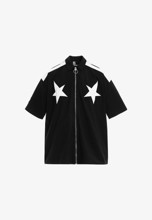 Persona joven que lleva una camiseta negra de manga corta y pantalones cortos con estrellas blancas, calcetines blancos y zapatos beige texturizados sin cordones, de pie sobre un fondo blanco.
