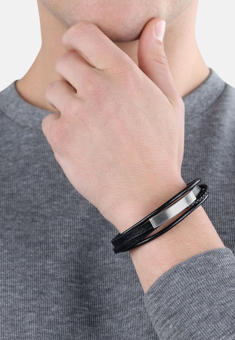 Pulsera de cuero negro que presenta tres trenzas con un acabado texturizado y un acento rectangular metálico. Lleva en una muñeca.