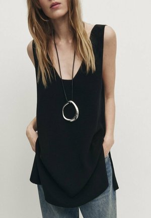 Femme portant un haut noir ample sans manches, un jean bleu, un long collier avec un grand pendentif argenté, mains dans les poches devant un fond uni.