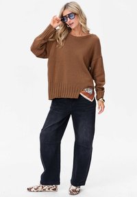 Bruin gebreid sweater met geribbelde zoom, ontspannen pasvorm, gecombineerd met donkerdenim wijde broek en gespikkelde instappers. Afgewerkt met een zonnebril.