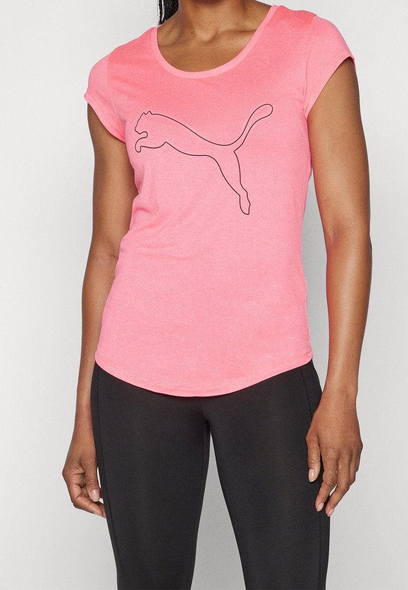 Kvinna klädd i en rosa kortärmad T-shirt med en svart omramad puma-logotyp och svarta leggings mot en enfärgad bakgrund.