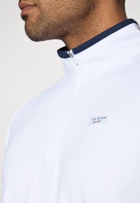 Balts sporta tops ar tumši zilu apkakli, ar ceturtdaļas rāvējslēdzēju un mazu zīmolu etiķeti uz krūtīm, gluda tekstūra un mūsdienīgs dizains.