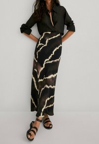 Black button-up shirt, patterned long skirt with dark and light abstract stripes, worn with black sandals, against a neutral background.