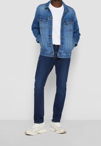 chaqueta vaquera, camiseta blanca, vaqueros azul oscuro y zapatillas de color claro. La chaqueta tiene bolsillos con botones y un corte holgado.