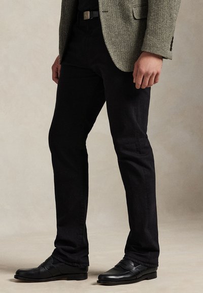 Man wearing black pants, black belt, black shoes, and a beige herringbone-patterned jacket standing against a plain background.