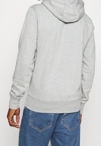 Sudadera gris con capucha, con puños y dobladillo acanalados. Llevada con jeans de mezclilla azul, mostrando un diseño simple y casual.