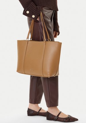 Persona sosteniendo un gran bolso tote de cuero marrón claro con asas dobles, vistiendo un suéter de punto marrón, pantalones de cuero marrón oscuro y zapatos planos puntiagudos oscuros.