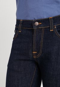 Mörkblå denimjeans med orange sömmar, med en framficka och en metallknapp som stängning i midjan. Slät textur.