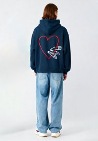 Felpa blu marine con cuore rosso ricamato e spille da balia bianche sul retro. Abbinata a jeans larghi blu chiaro e sneakers bianche.