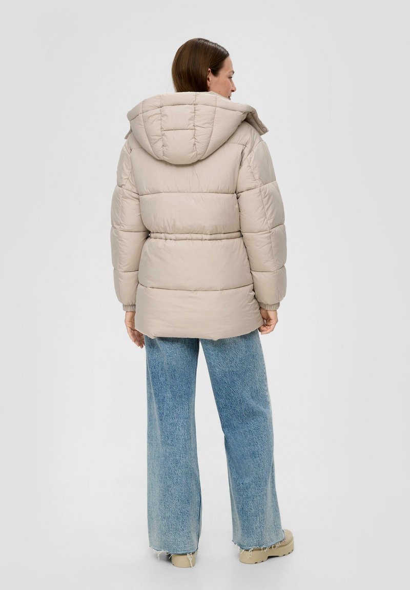 Wattierte Jacke in hellbeige mit Kapuze, mit horizontalem Steppmuster und elastischen Bündchen, kombiniert mit weit geschnittenen blauen Jeans.