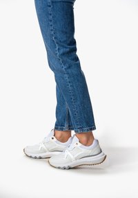White athletic shoes with mesh and synthetic materials, featuring textured soles and gold accents. Paired with blue denim jeans.