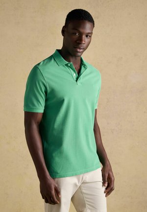 Young man wearing a green short-sleeve polo shirt and light beige pants standing against a plain beige background.