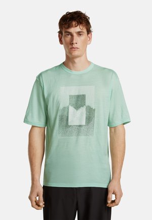 Homme portant un T-shirt vert clair à manches courtes avec un motif géométrique rectangulaire dans des nuances de vert plus foncé à l'avant.