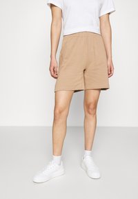 Breda bomullsshorts i beige med elastisk midja och sidfickor, kombinerade med vita sneakers och ankelsockor, på en neutral bakgrund.