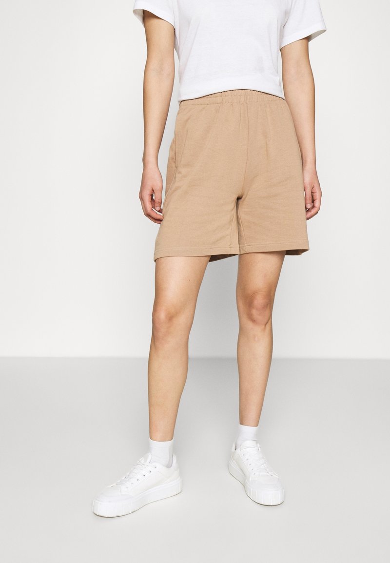 Breda bomullsshorts i beige med elastisk midja och sidfickor, kombinerade med vita sneakers och ankelsockor, på en neutral bakgrund.