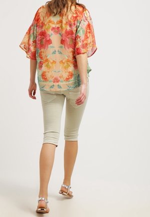 Blusa floreale trasparente con fiori arancioni e rosa vivaci, vestibilità comoda, indossata su pantaloni capri beige chiaro, abbinata a sandali argento.