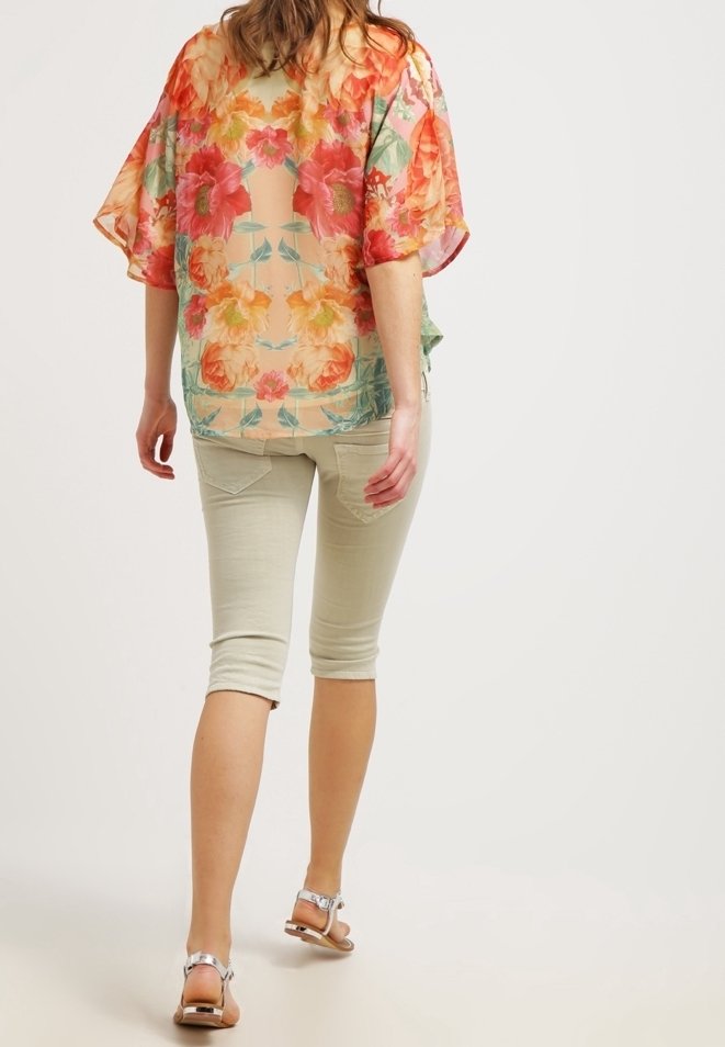 Hihna bluzė su spalvingais oranžiniais ir rožiniais gėlėmis, laisvas kirpimas, uždėta ant šviesių smėlio spalvos capri kelnių, derinama su sidabriniais šlepetėmis.