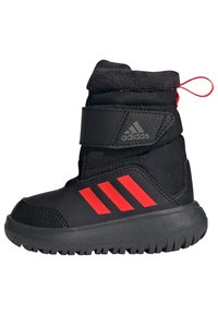 Zwarte winterlaars met een grijs Adidas-logo, rode strepen aan de zijkant en een stevige textuur rubberen zool. Voorzien van een klittenbandsluiting en een treklipje.