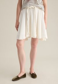 Falda de patinadora blanca con tejido texturizado, cinturilla elástica y detalle de cordones trenzados; combinada con zapatos planos en verde olivo.