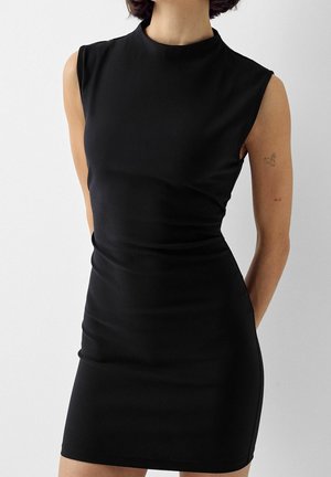 Kobieta w przylegającej, czarnej sukience bez rękawów z wysokim dekoltem, stojąca na tle jednolitego jasnego tła, ukazująca górną część tułowia i ramiona.