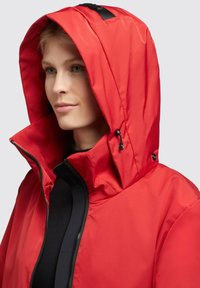 Chaqueta impermeable roja con cuello alto y capucha ajustable, con un zipper negro y una marca discreta a lo largo de los bordes. Textura suave.