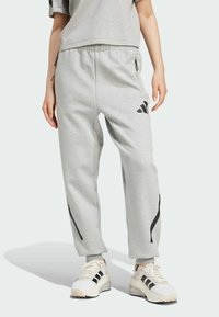 Pantaloni della tuta grigio chiaro con vestibilità rilassata, vita elasticizzata, cerniere laterali e strisce nere lungo i lati, abbinati a dettagli bianchi delle sneakers.