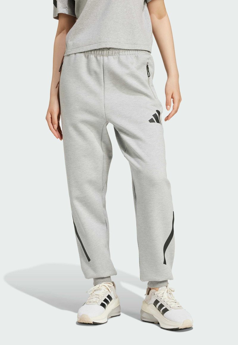 Pantaloni della tuta grigio chiaro con vestibilità rilassata, vita elasticizzata, cerniere laterali e strisce nere lungo i lati, abbinati a dettagli bianchi delle sneakers.