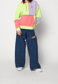 Colorful hooded sweatshirt with green, purple, pink sections; features a logo and drawstring. Wide-leg denim pants with cutouts. White sneakers.