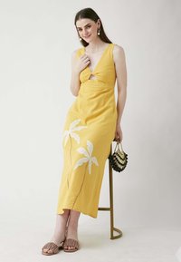 Gelbes Leinenkleid mit V-Ausschnitt und Schlüsselloch-Detail, verziert mit weißer Palmenblatt-Stickerei. Dazu werden beigen Sandalen und einer schwarzen Handtasche getragen.