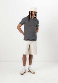 Striped gray T-shirt, white cotton shorts, and beige bucket hat. Light sneakers with brown soles. Model poses with hands on hips.