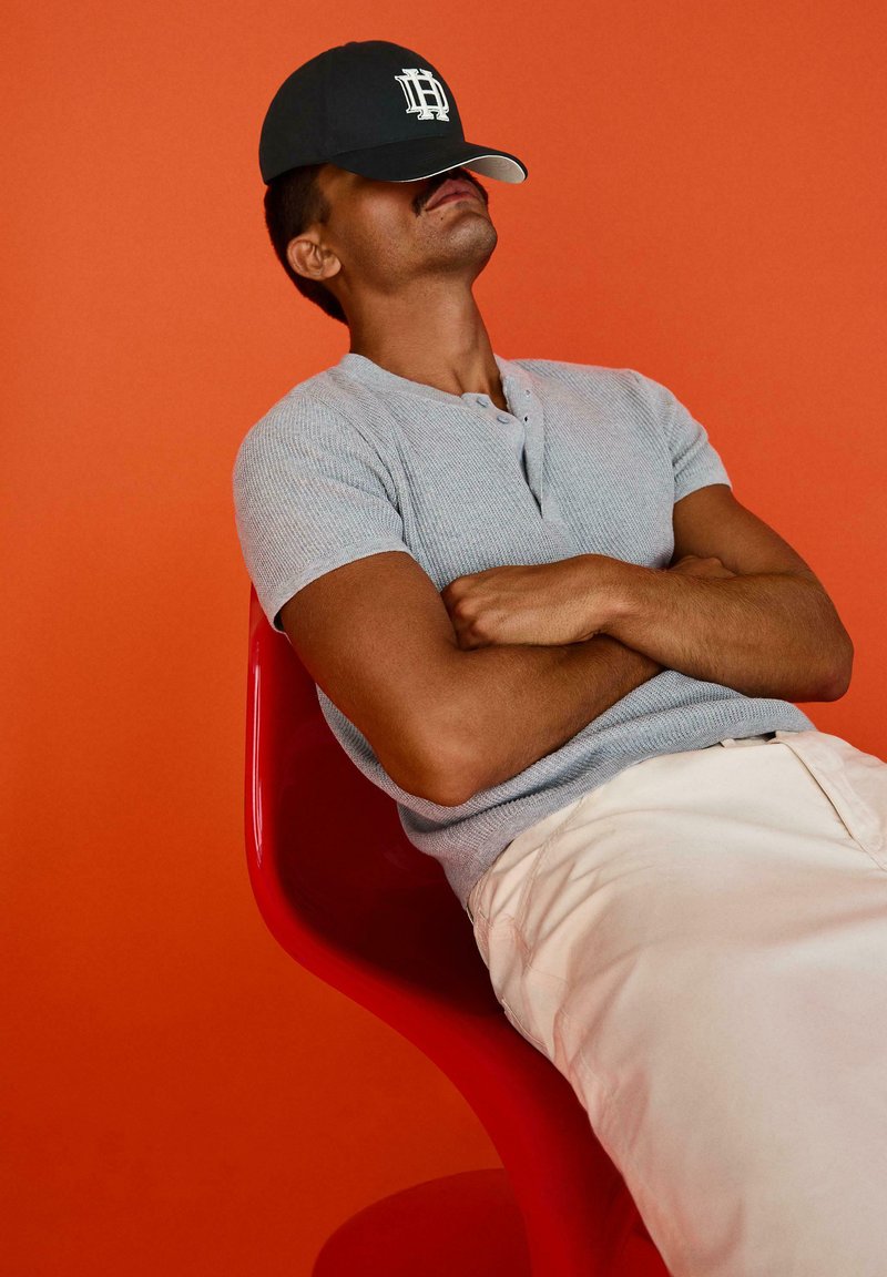 Homme portant une chemise grise et une casquette noire inclinée sur les yeux, assis en arrière les bras croisés sur une chaise rouge sur fond orange.