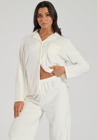 Witte pyjama set gemaakt van zachte stof; bestaat uit een button-up shirt met een borstzak en broek met elastische taille. Soepele textuur en een comfortabele pasvorm.