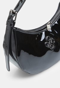 Sac à main en cuir verni noir brillant avec bandoulière réglable, fermeture éclair argentée et emblème métallique logo sur le devant.