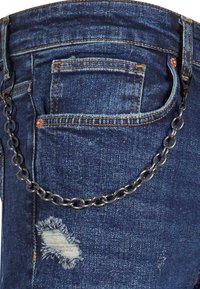 Gros plan sur un jean en denim bleu avec une chaîne métallique attachée de la boucle de ceinture à la poche avant et une déchirure effilochée visible près de la cuisse.