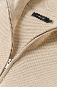 Beige zipzáras pulóver texturált kötésmintával, ezüst szippantóval, és egy fekete címkével, rajta a "Matinique" márkanevével és "L" mérettel.