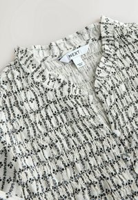 Blouse blanche texturée avec imprimé floral noir et col à volants, dotée d'une étiquette de marque "NEXT" et d'une étiquette « Fabriqué en Inde ».